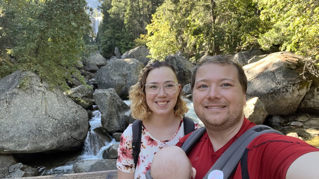 Family of mom, dad and baby take a selfie on the footbridge of the Yosemite mist trail with vernal falls in the background