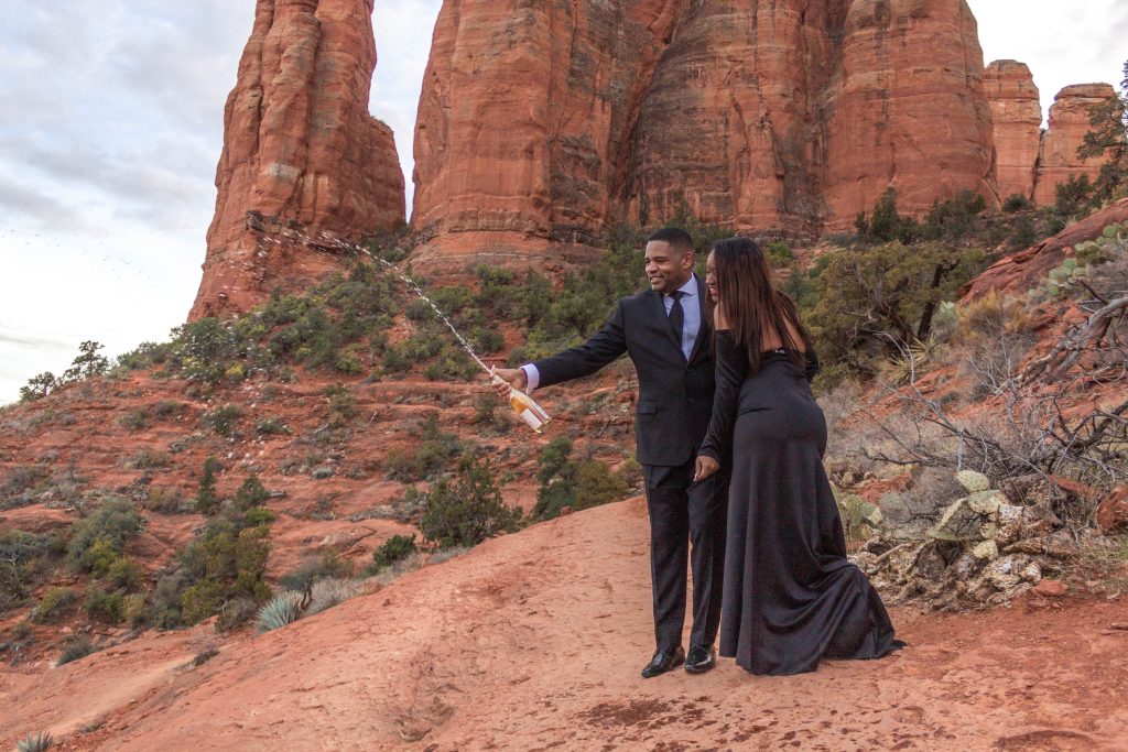 Couple standing on the trail during a cathedral rock engagement session. They are dressed in all black shaking a champagne bottle